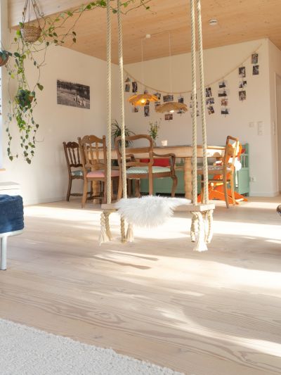 Espace de vie lumineux avec sol en bois, balançoire avec housse en fourrure blanche au centre, table à manger avec chaises à l’arrière, escalier à droite et plantes suspendues au plafond, avec parquet en douglas.