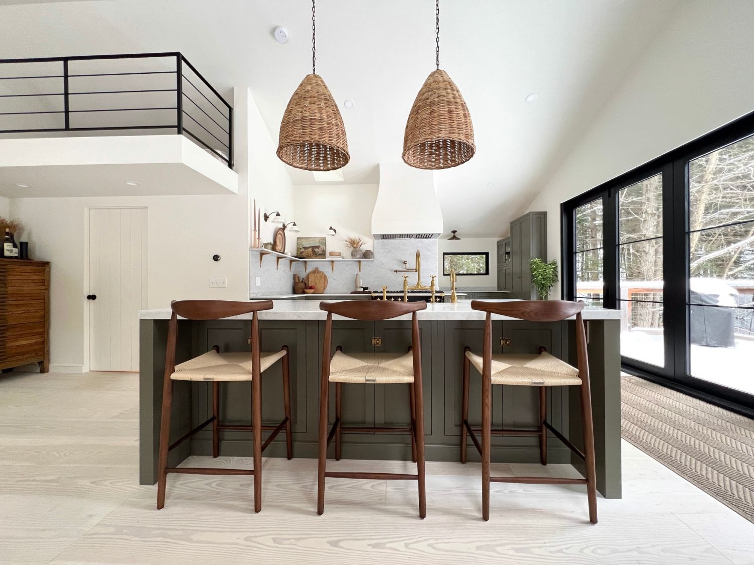 Modern kitchen with kitchen island, three wooden chairs with light seats, and two woven pendant lights above the island, large windows with views of a snowy landscape.