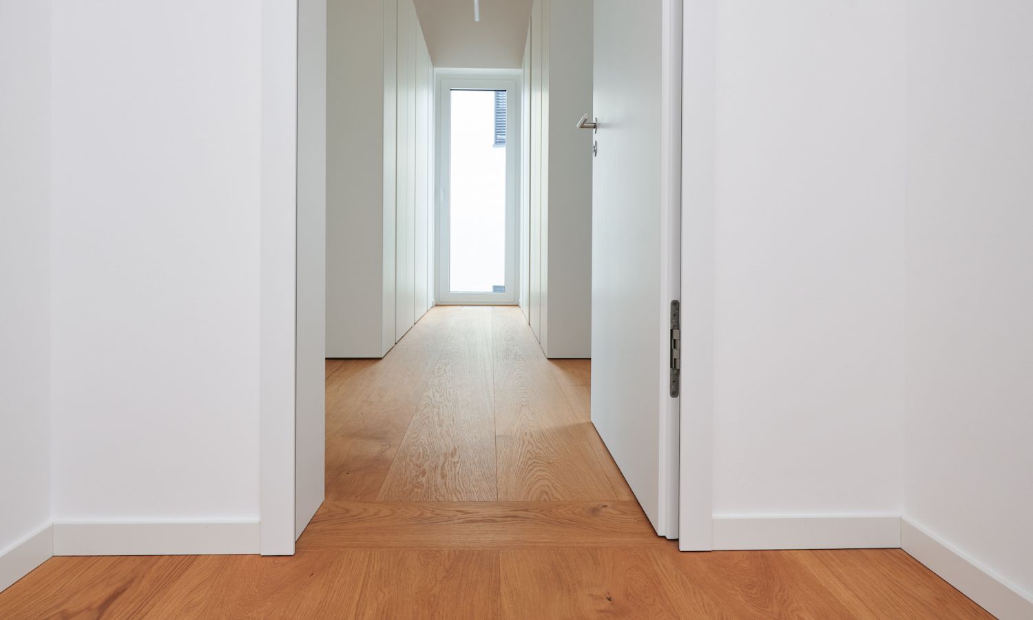 View through a white hallway with an open white door and oak floorboards, a window with daylight at the end.