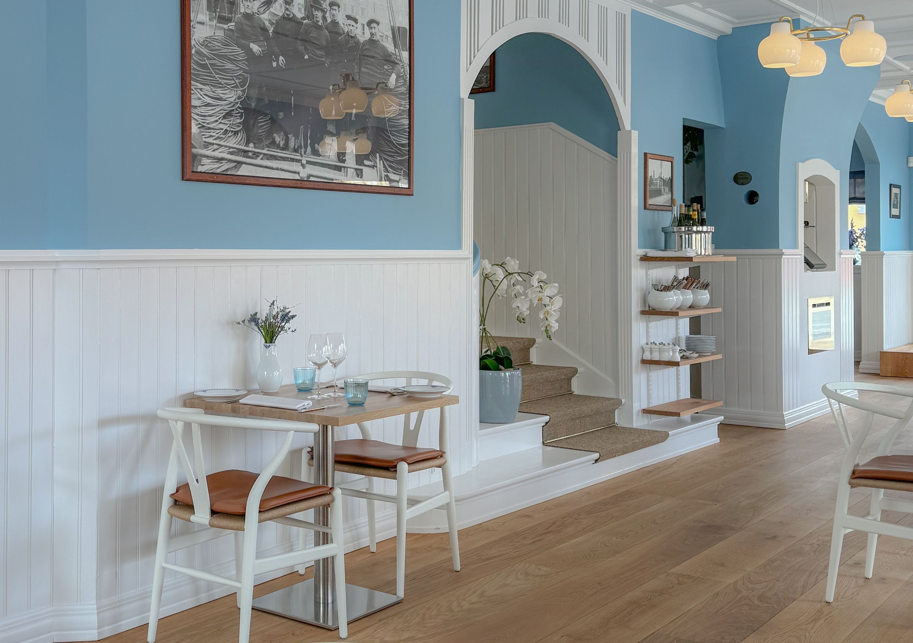 Salle lumineuse bleu clair avec lambris blancs, parquet en chene, deux tables avec chaises blanches et coussins bruns, escalier et etagere murale avec vaisselle.