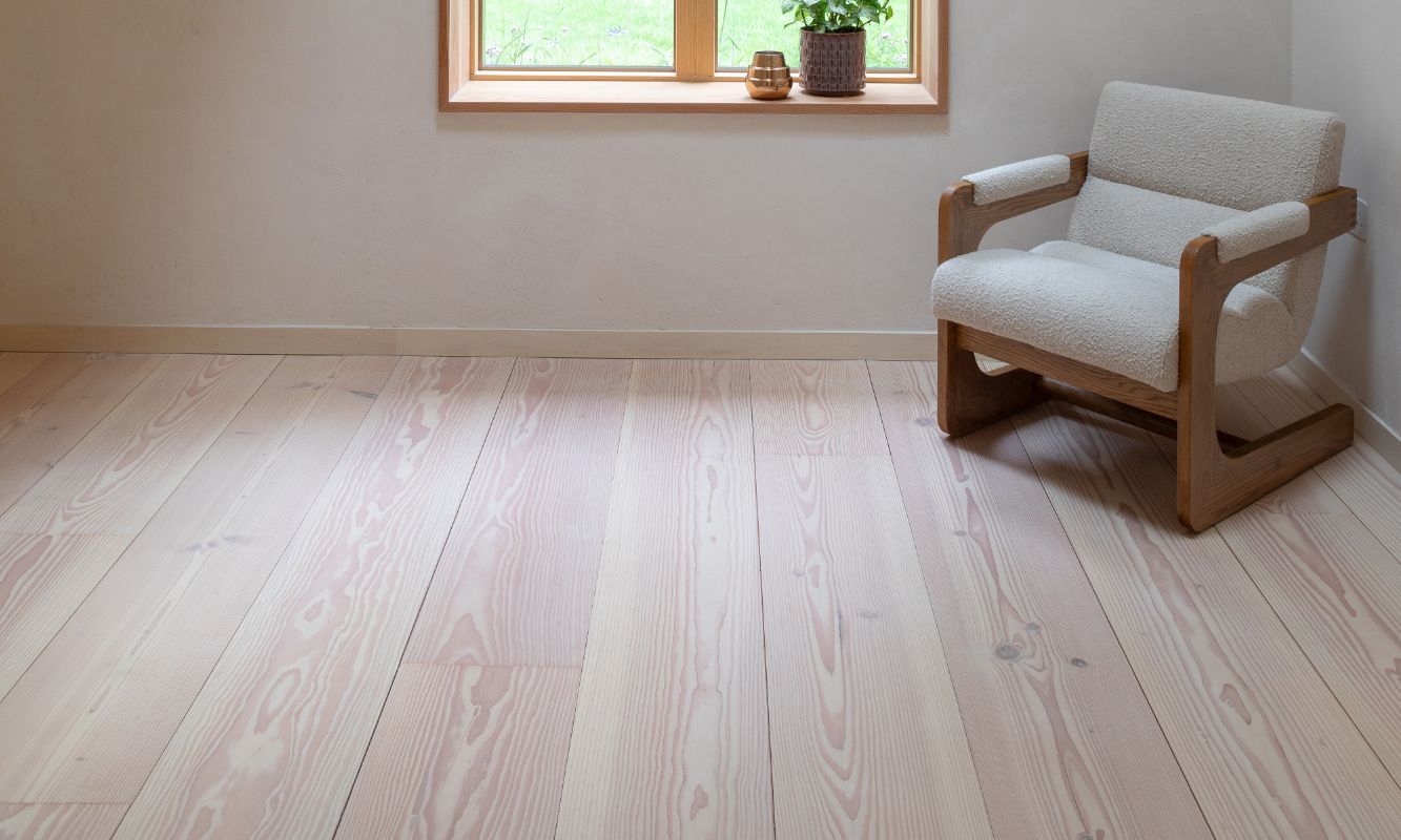 Blick in ein Zimmer und darüber hinaus in den Garten. Im Zimmer befindet sich ein einzelner Sessel in der Ecke, ein heller Holzboden mit markanter Maserung und zwei schlichte Wandleuchten.