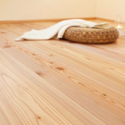 Gros plan d’un parquet en meleze huile clair, avec un panier tresse, une couverture blanche et une bougie en arriere-plan.
