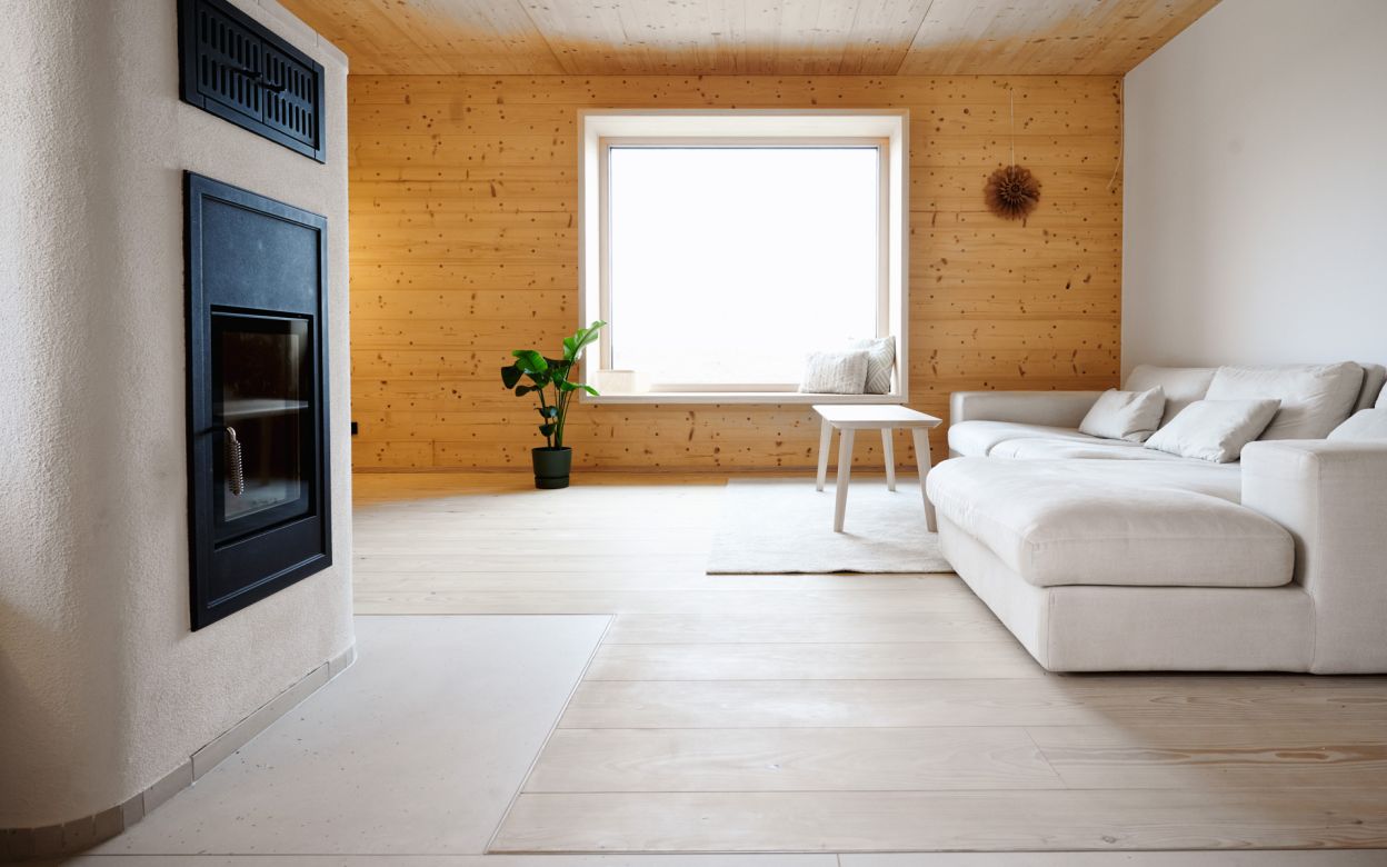 Helles Wohnzimmer mit weissem Ecksofa, kleinem Holztisch, gruener Topfpflanze und Kamin mit schwarzer Tuer.