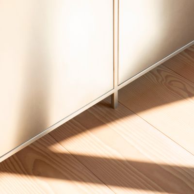 Détail d’une cuisine en métal inoxydable et d’un sol en planches de Douglas. La lumière douce tombe de la droite sur la façade de la cuisine et sur la partie du sol.