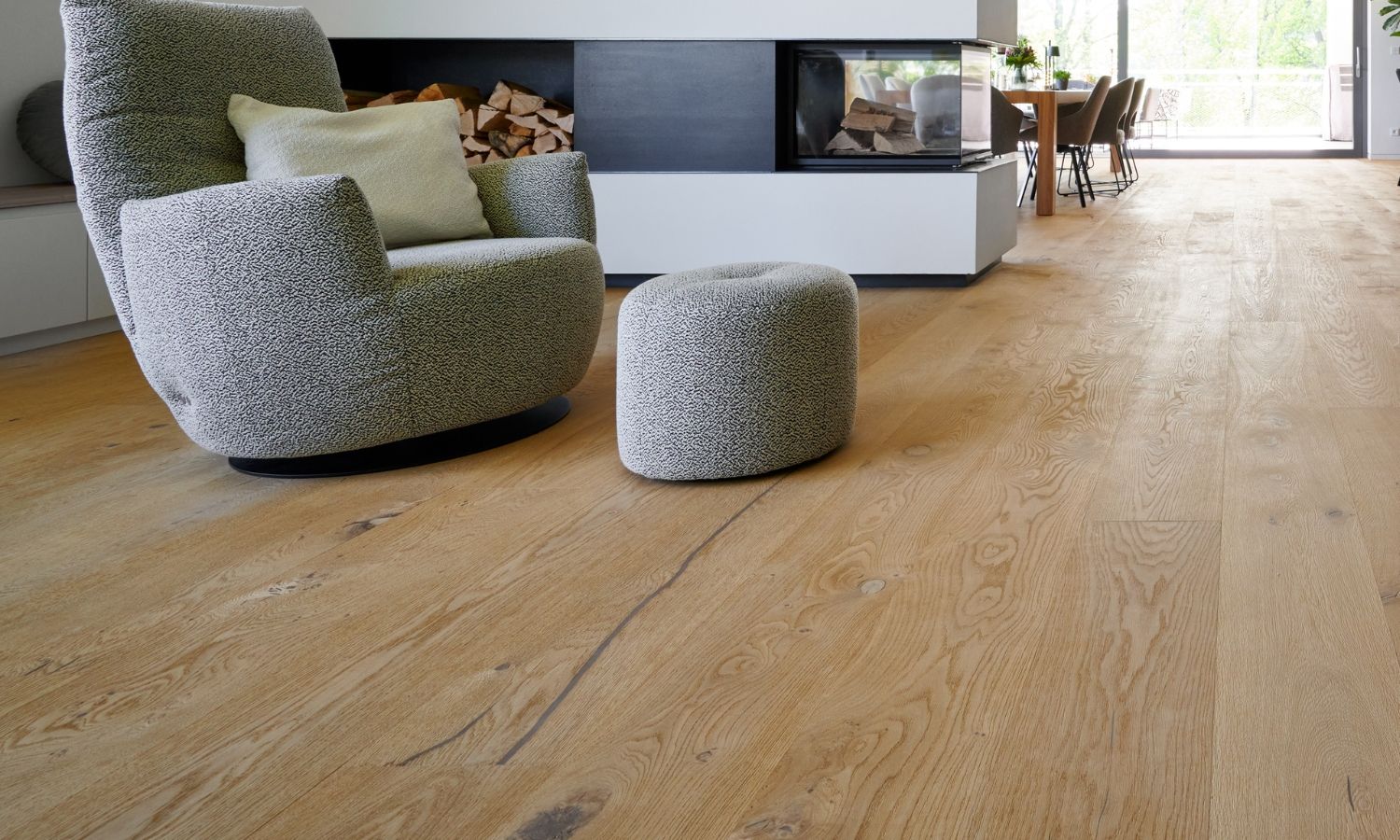 Fauteuil rembourre gris clair avec pouf assorti sur un sol en chene devant une cheminee moderne avec rangement pour le bois.