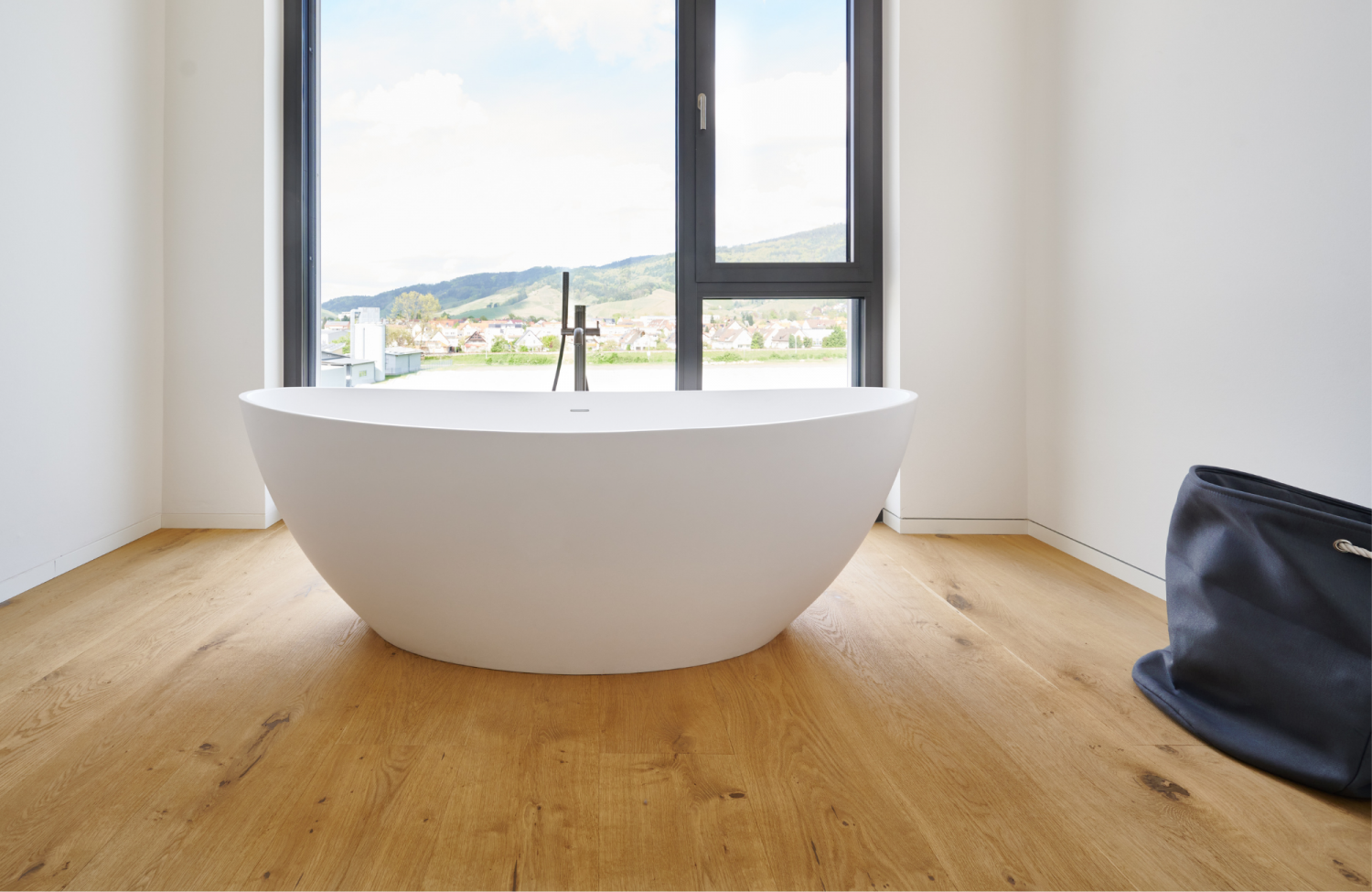 Freistehende weisse Badewanne vor grossem Fenster mit Blick auf Huegel, Eiche Holzboden, schwarze Tasche rechts.