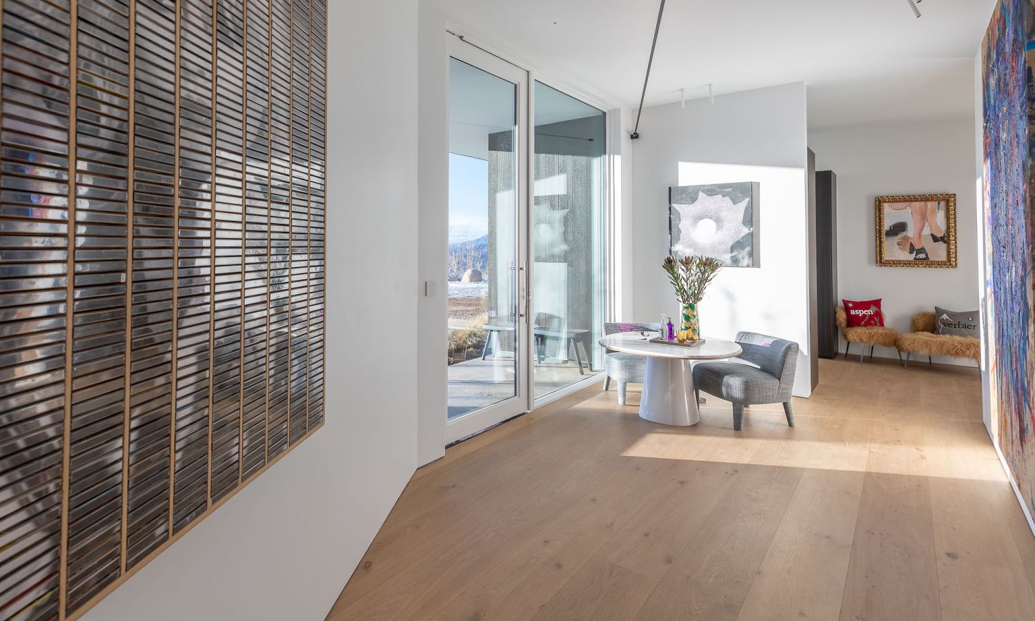 Piece lumineuse et moderne avec sol en bois de chene, grande oeuvre murale en lattes de bois a gauche, table ronde avec chaises et fleurs au centre, autre tableau sur le mur de droite et canape en arriere-plan.