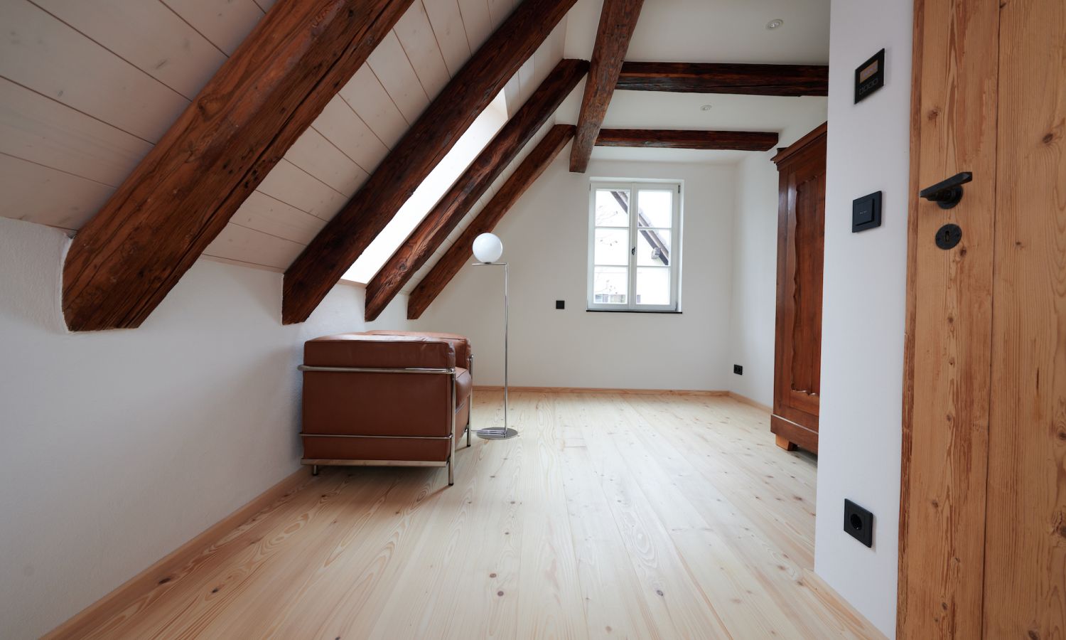 Chambre mansardee avec poutres apparentes, canapé en cuir brun, lampadaire et porte en bois à côté de la fenêtre avec murs blancs et parquet en sapin blanc.