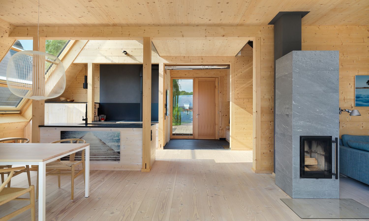 Espace de vie ouvert avec murs et sol en bois de douglas, cheminée moderne grise, ensemble de salle à manger blanc et cuisine avec façades noires.