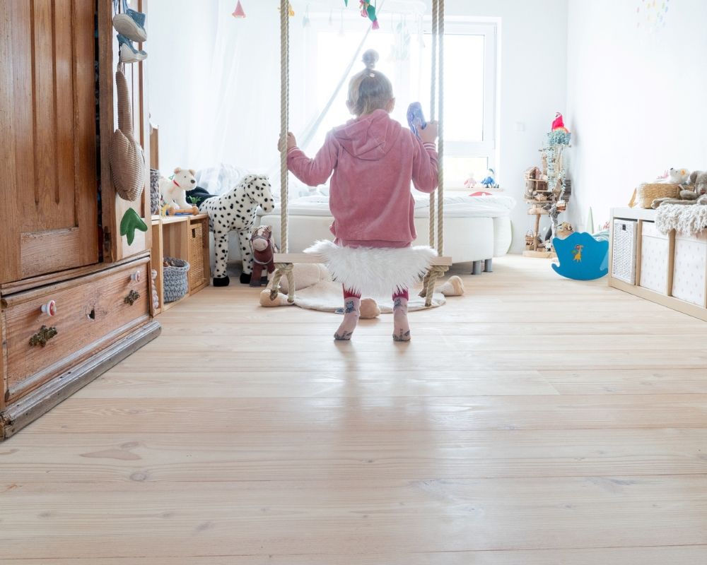 Kind mit rosa Kapuzenjacke und weißem Tüllrock schaukelt in einem hellen Kinderzimmer mit Douglasie Holzfußboden und Spielzeug.