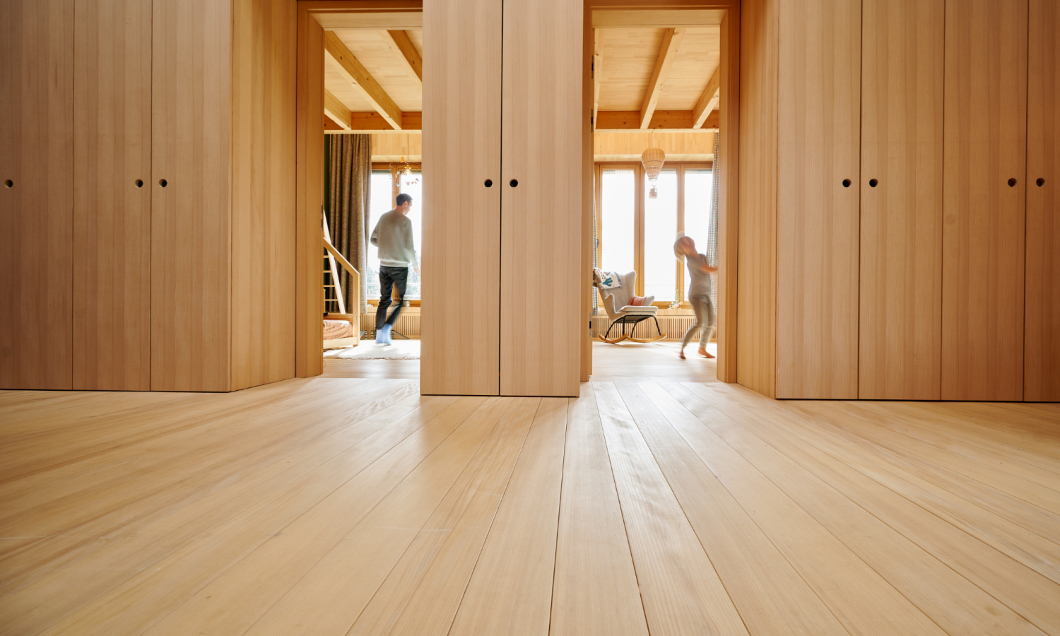 Blick auf hellen Holzboden und zwei offene Tueren mit Holzvertafelung, zwei Personen in Bewegung im Hintergrund.