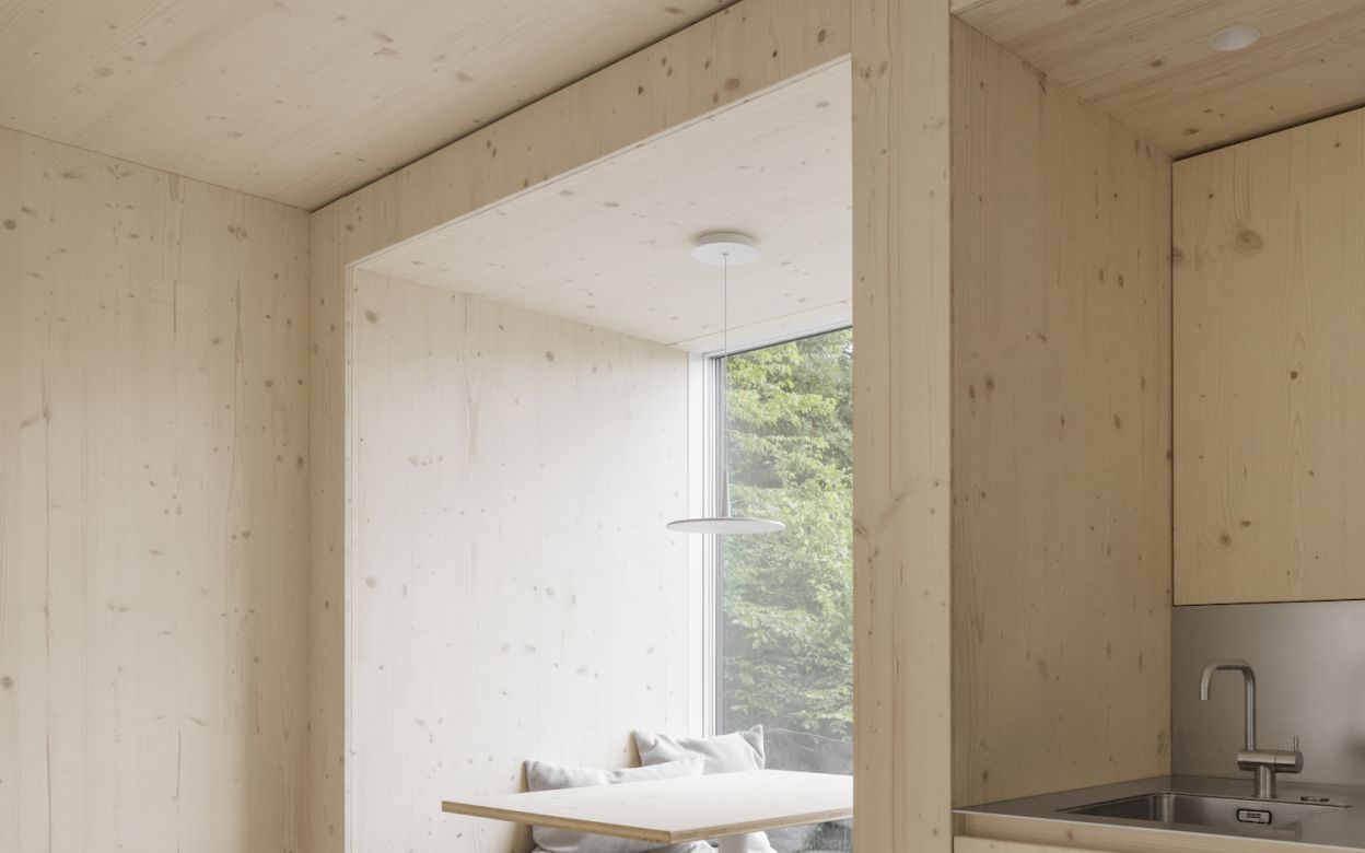 Moderner Innenraum mit hellen Holzflaechen, integrierter Sitzbank mit grauen Polstern und kleinem Esstisch vor Fenster mit Blick ins Grueene.