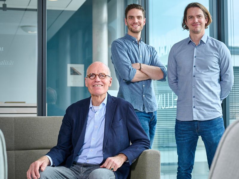 Famille von Saucken dans un bureau moderne, une personne assise dans un fauteuil, deux debout derrière devant de grandes fenetres.