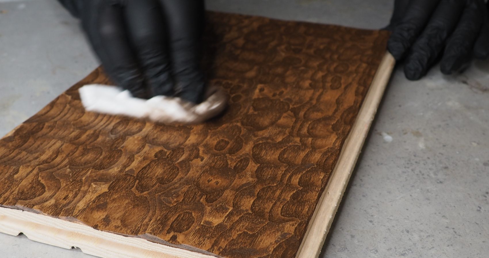 Eine Hand streicht mit einem Tuch über ein Stück einer Wandpaneele aus Holz und trägt dunkles Öl auf.
