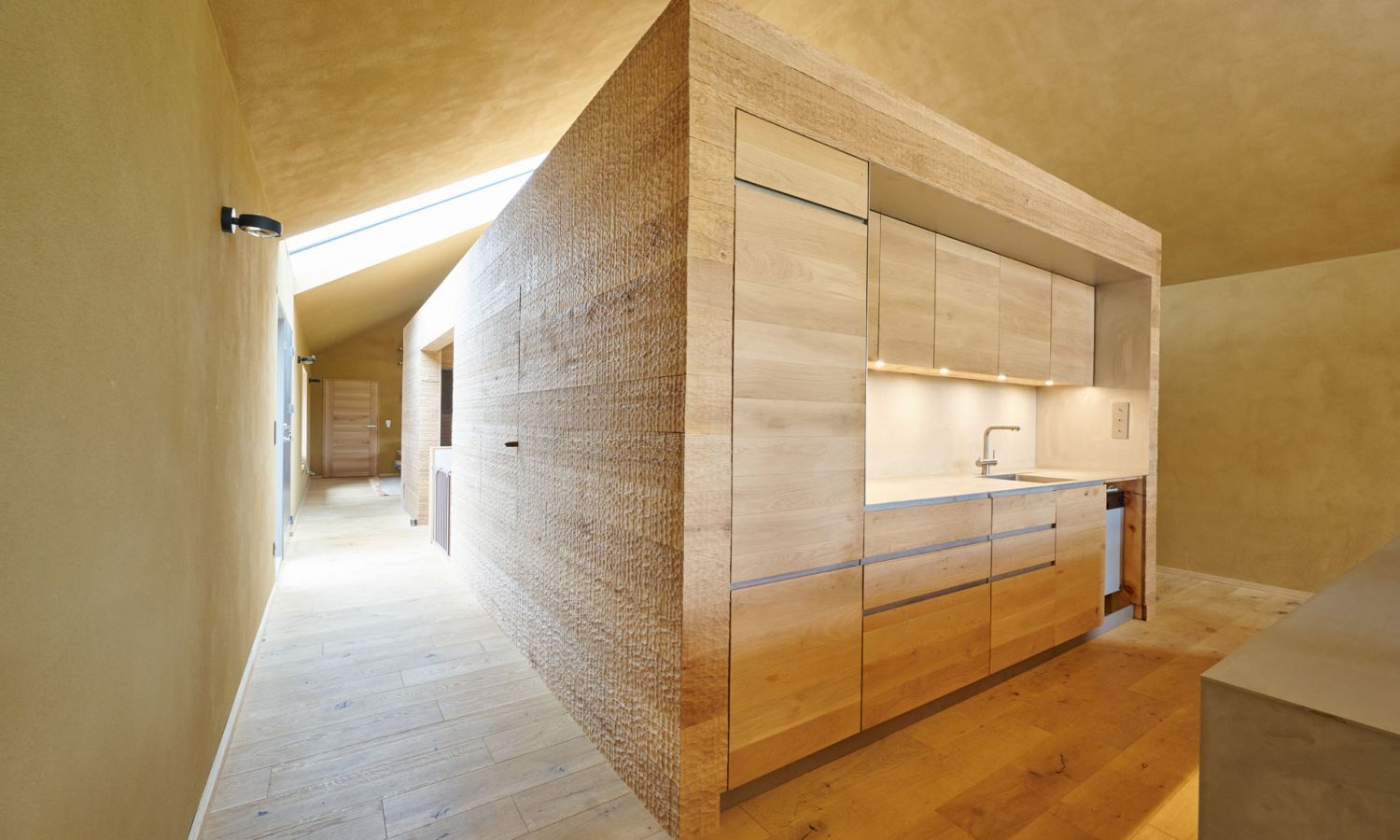 View of a room featuring a large square interior cube in the center. The entire cube is clad in wood, with a kitchen unit integrated at the front. The walls are plastered with clay.