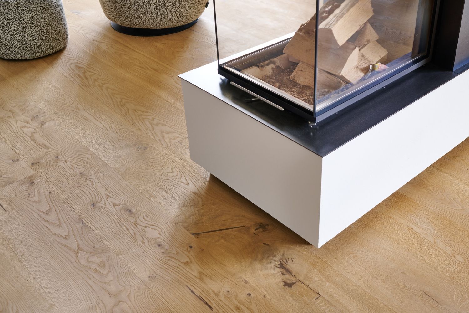 Moderner, rechteckiger Kamin mit Glasfront und Holzscheiten auf Eiche Holzboden, im Hintergrund zwei runde Sitzhocker.