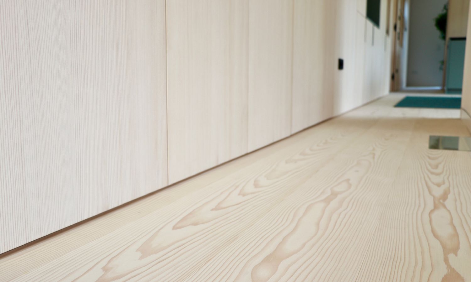 Vue sur un parquet en sapin blanc avec veinures visibles et un mur avec des portes d’armoires claires dans un couloir.