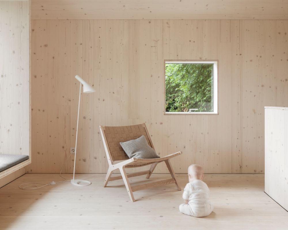 Enfant en body blanc assis sur le sol en bois de sapin blanc devant une chaise en bois avec un coussin gris, à côté d’une lampe sur pied blanche, avec une fenêtre en arrière-plan donnant sur des plantes vertes.