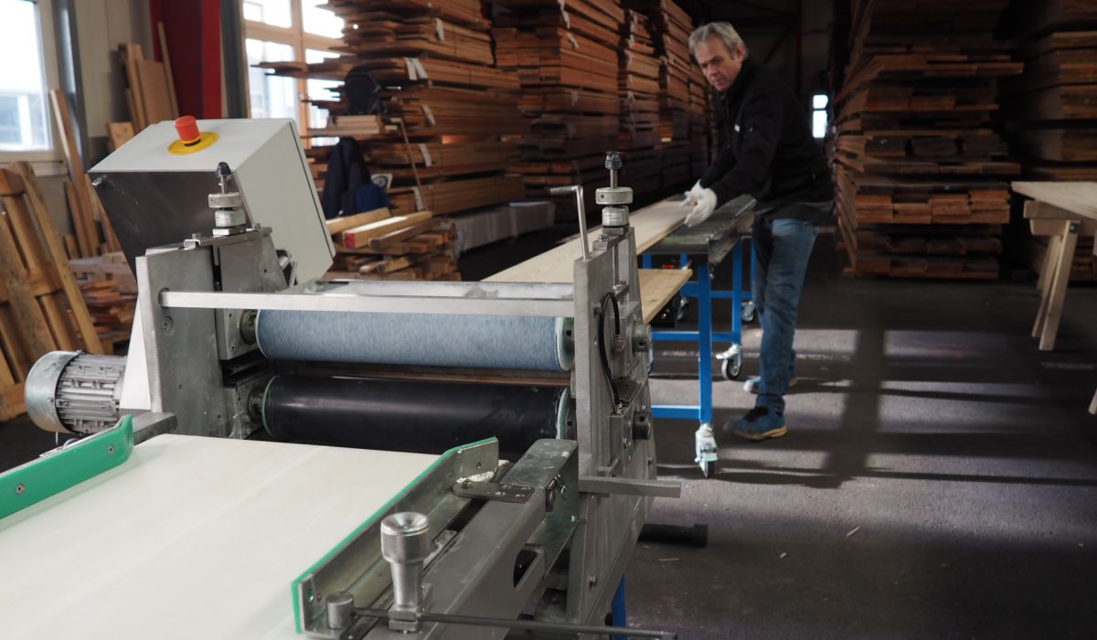 Person in dark clothing operates a large woodworking machine in a workshop with stacked wood in the background.