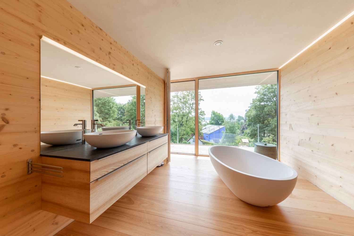 Modernes Badezimmer mit freistehender ovaler Badewanne, zwei runden Waschbecken auf Holzwaschtisch und grossem Fenster mit Blick ins Gruene.