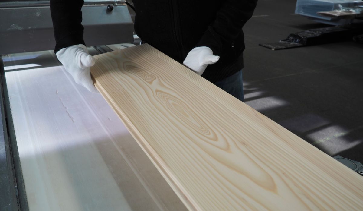 Two hands wearing white gloves hold a light wooden board with visible grain in a workshop.