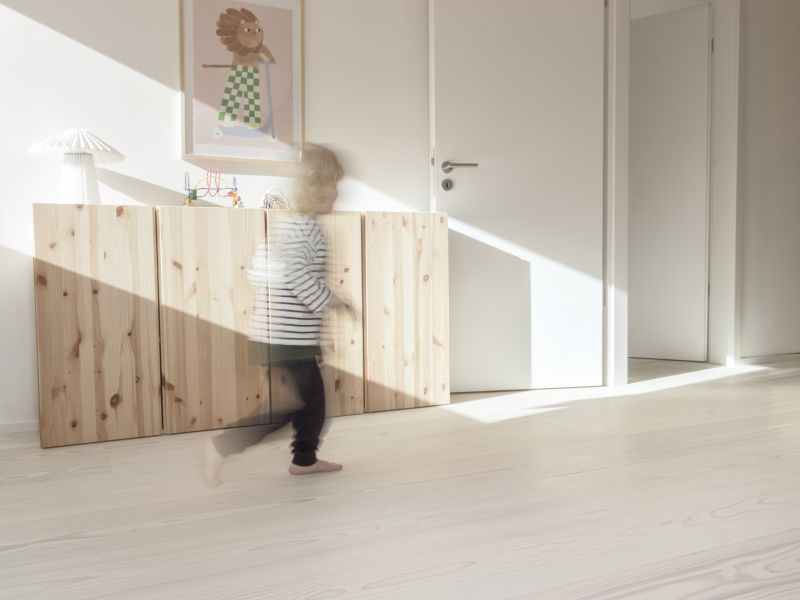 Kind läuft barfuß im Kinderzimmer; Schrank aus Holz mit einer Lampe und einem Bild mit einem Löwen im Hintergrund; Heller Holzboden; weiße Wände und eine weiße offene Tür