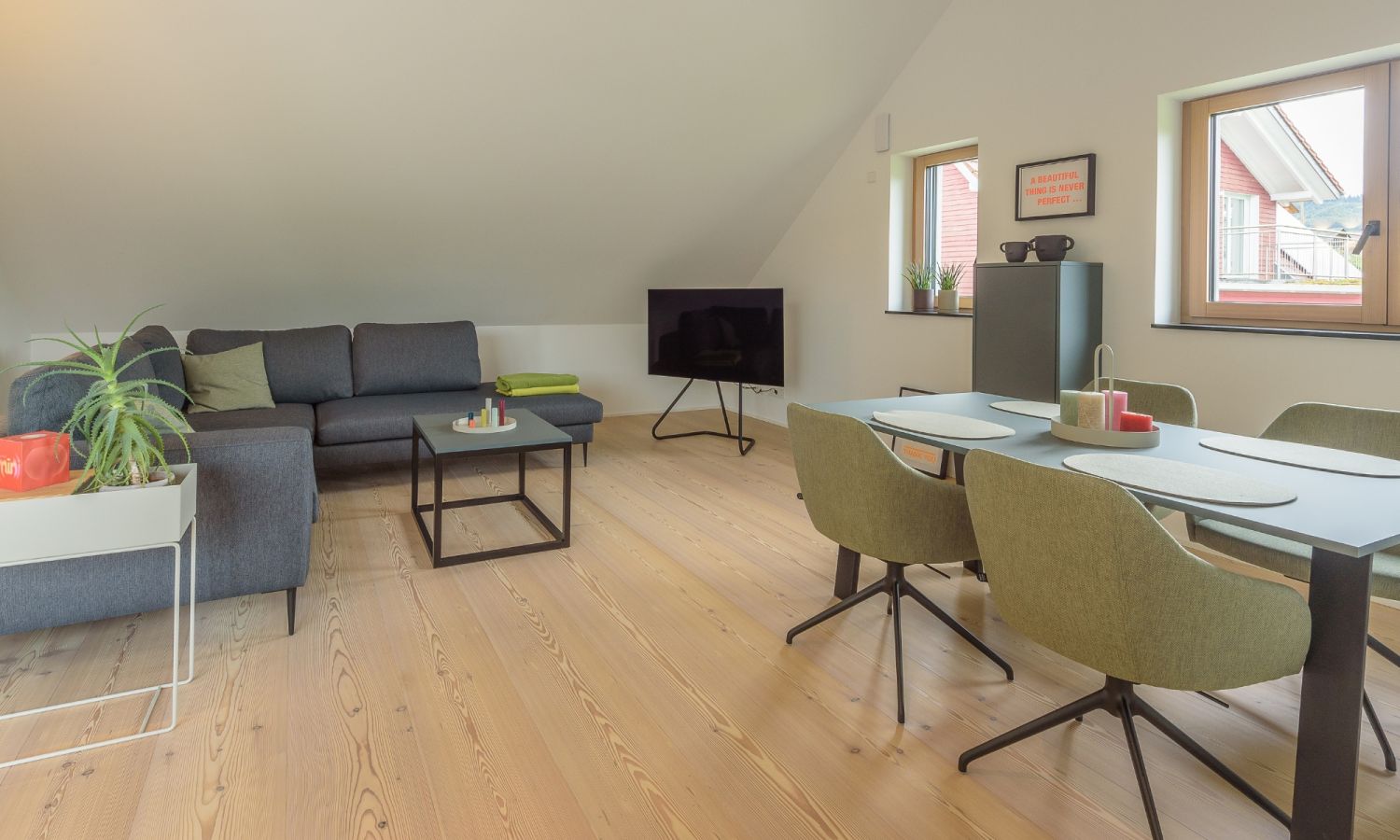 Salon avec canape d’angle gris, table basse noire, table a manger verte avec quatre chaises, televiseur sur meuble et deux fenetres.