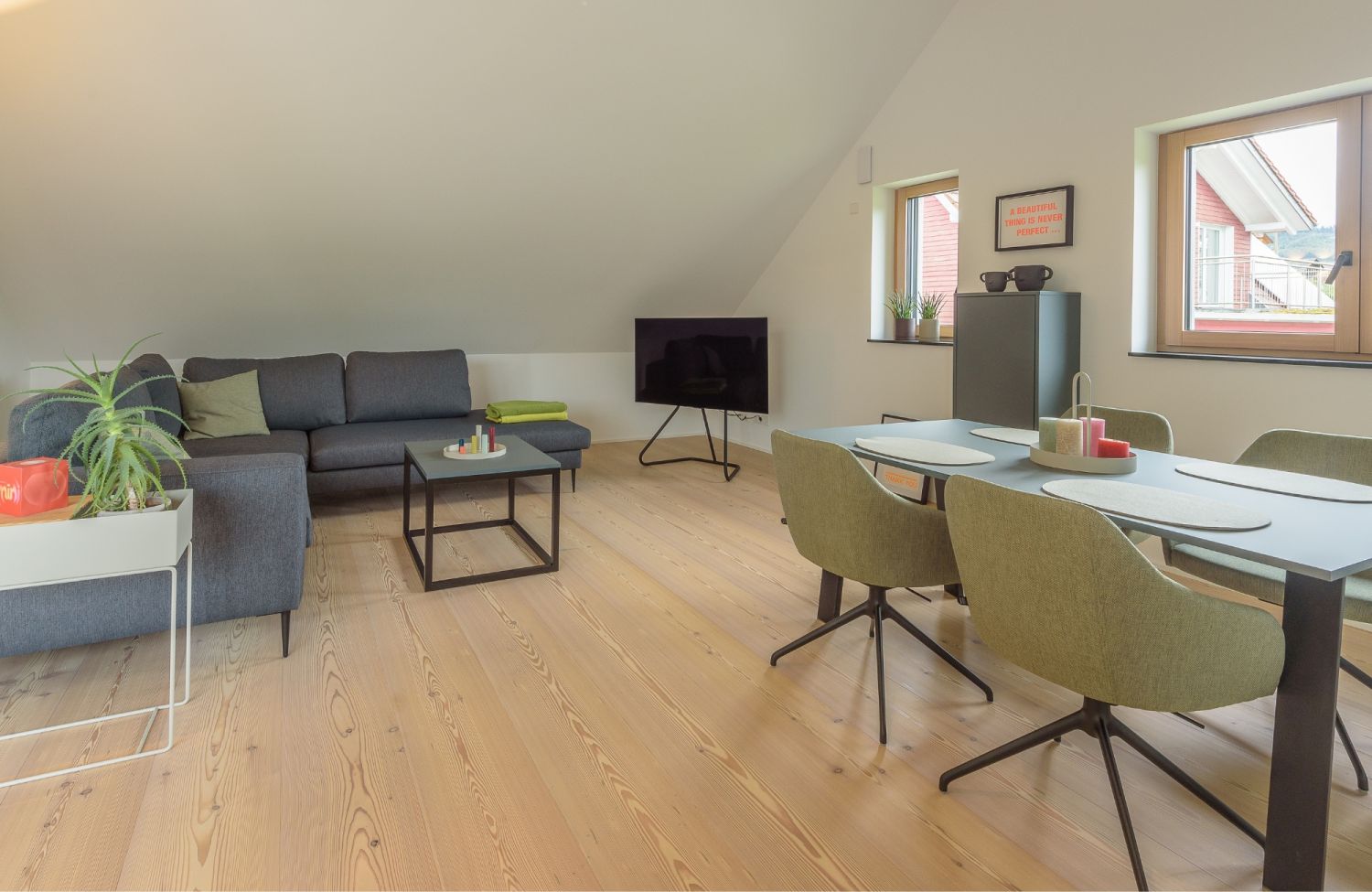 Wohnzimmer mit grauem Ecksofa, schwarzem Couchtisch, gruenem Esstisch mit vier Stuehlen, Fernseher auf Staender und zwei Fenstern.