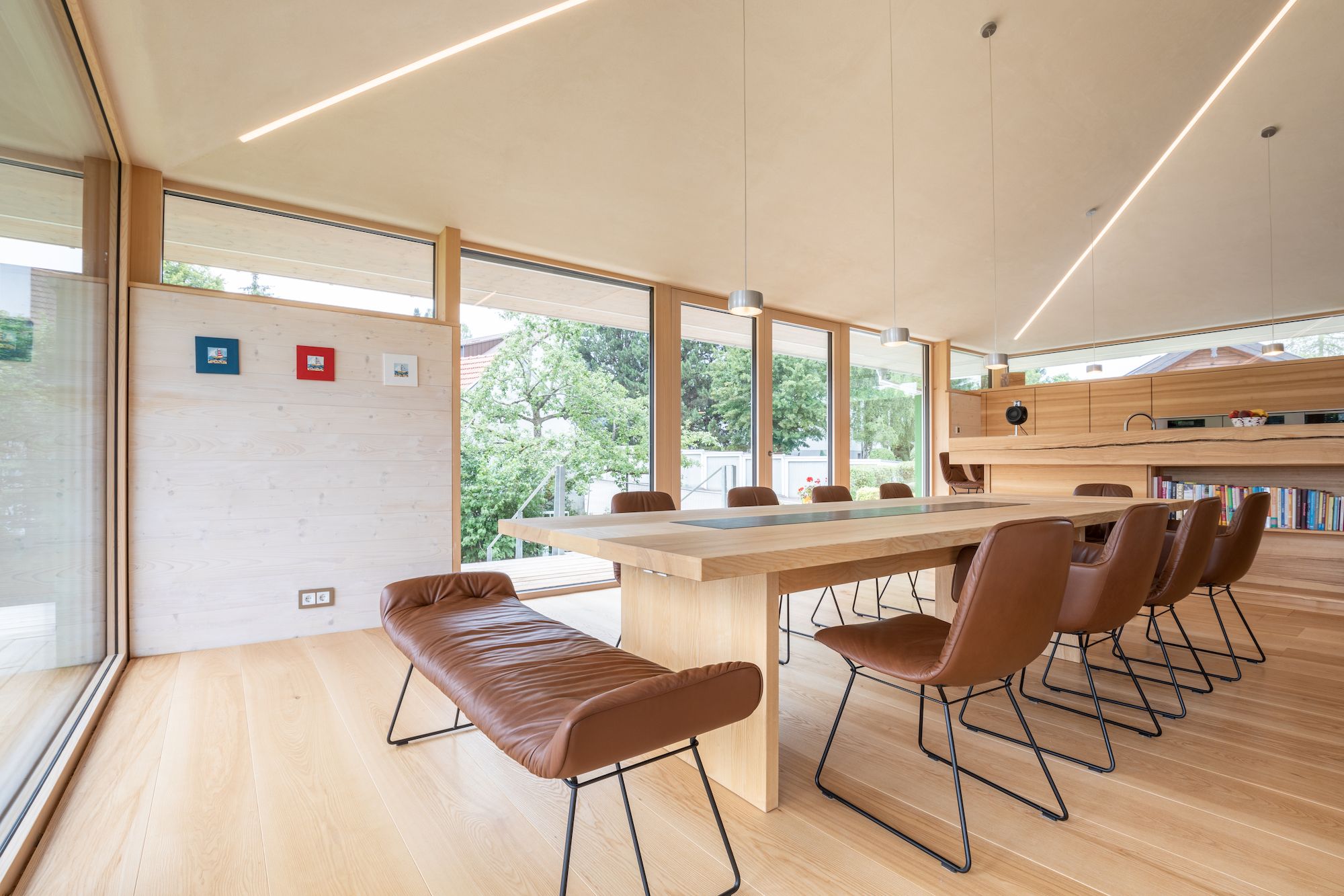 Moderner Essbereich mit langem Holz Tisch, braunen Lederstuehlen und Bank, grossen Fenstern mit Blick auf gruene Baeume.