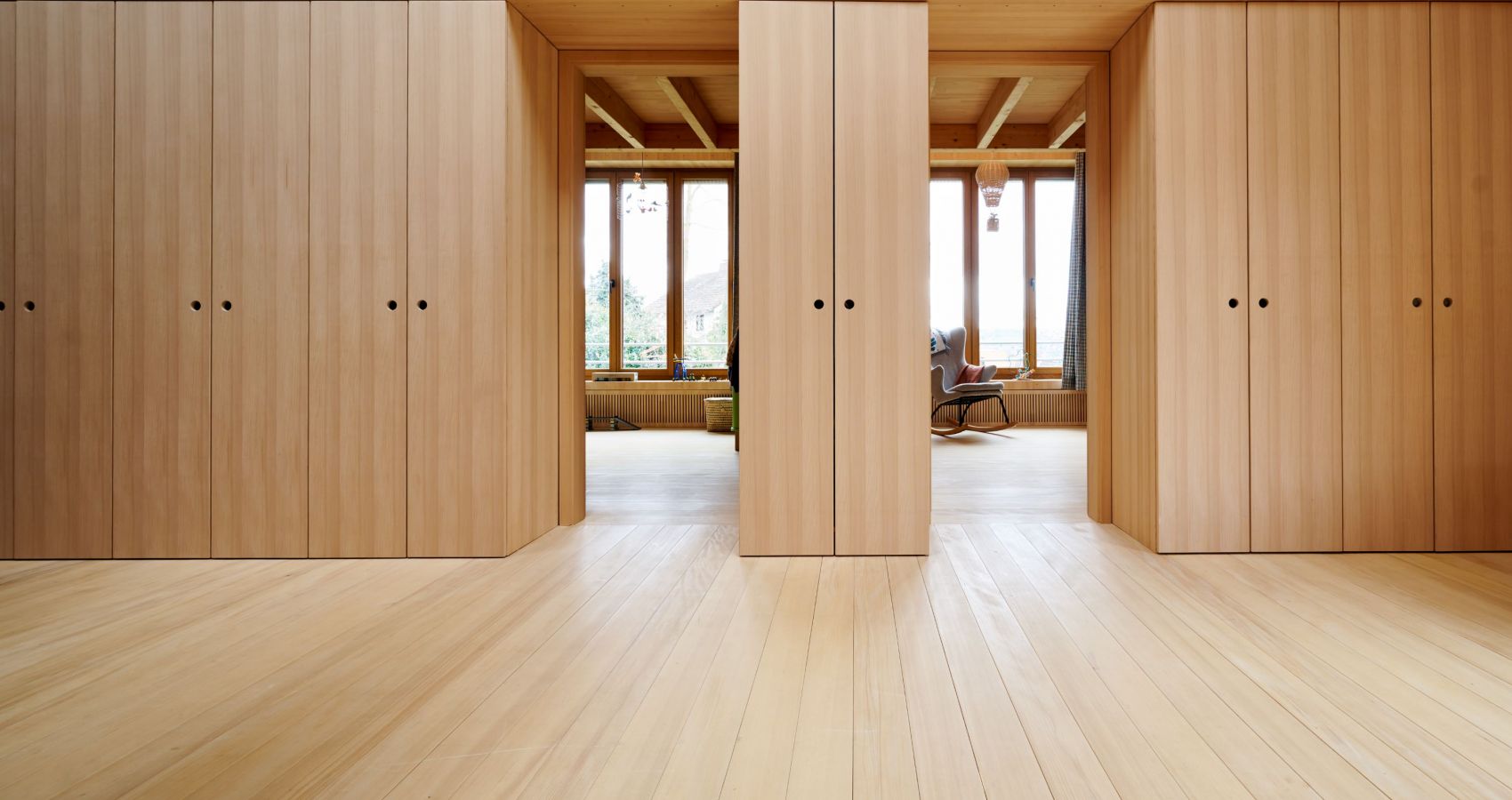 Grosser Raum mit Weisstanne Holzboeden und mehreren hohen, schlichten Holzschraenken mit schwarzen Griffen, Durchgang zu einem Sitzbereich mit grossen Fenstern.