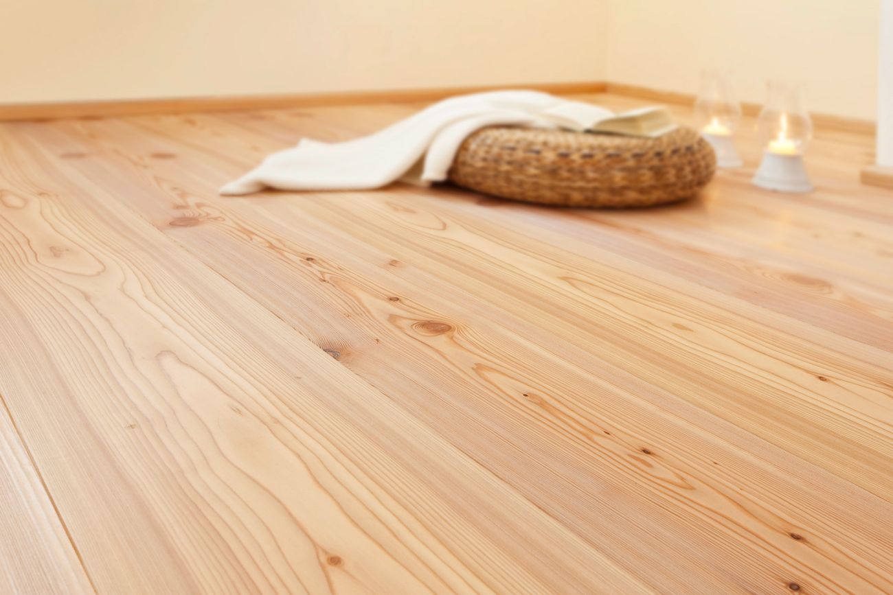 Close-up of a light oiled larch wood floor with a woven basket, a white blanket, and a candle in the background.
