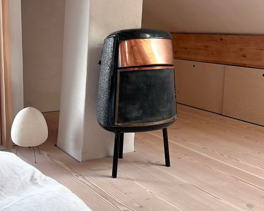 Ein kleiner alter Holzofen aus Douglasie und Messing steht vor einer Säule aus verputztem Kalkstein. Daneben befindet sich eine kleine Lampe, und der Boden besteht aus Douglasie-Holz mit hellem, leicht rosafarbenem Ton
