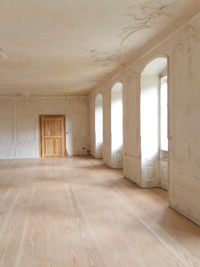 Ancienne salle à manger d’un château. À droite, de grandes fenêtres en arc laissent entrer la lumière. Au fond de la pièce, une porte en bois est visible. Le sol est recouvert de planches de sapin de Douglas de la longueur de la pièce.
