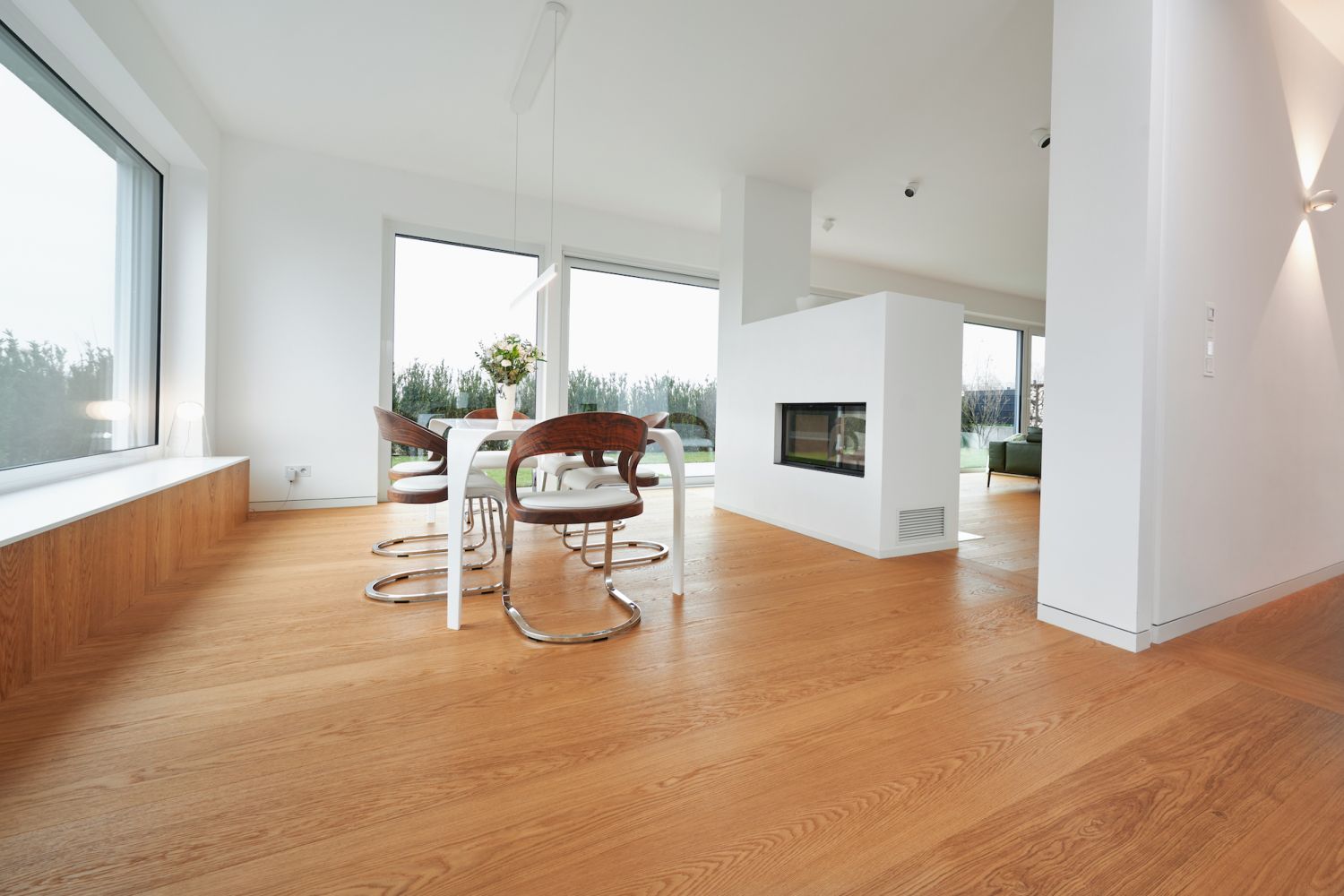 Moderner Essbereich mit weissem Tisch, braunen Stuehlen mit Metallgestell und mittig eingebautem Kamin in hellem Raum mit grossen Fenstern