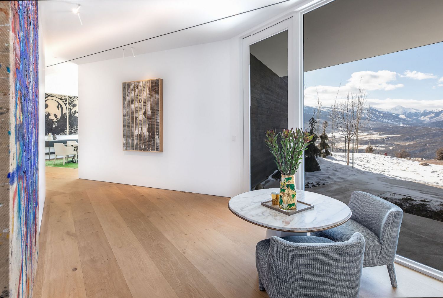 Modern living area with oak wood flooring, round marble coffee table, two grey armchairs and large window with mountain view.