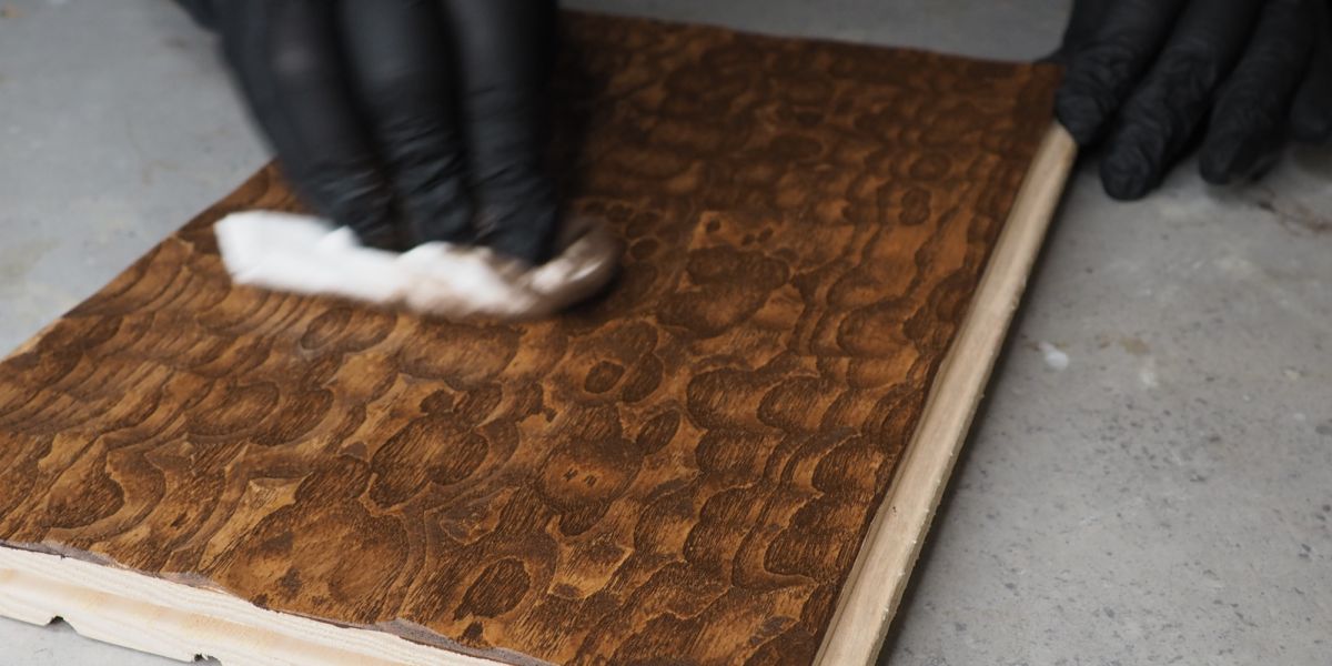 Eine Hand streicht mit einem Tuch über ein Stück einer Wandpaneele aus Holz und trägt dunkles Öl auf.
