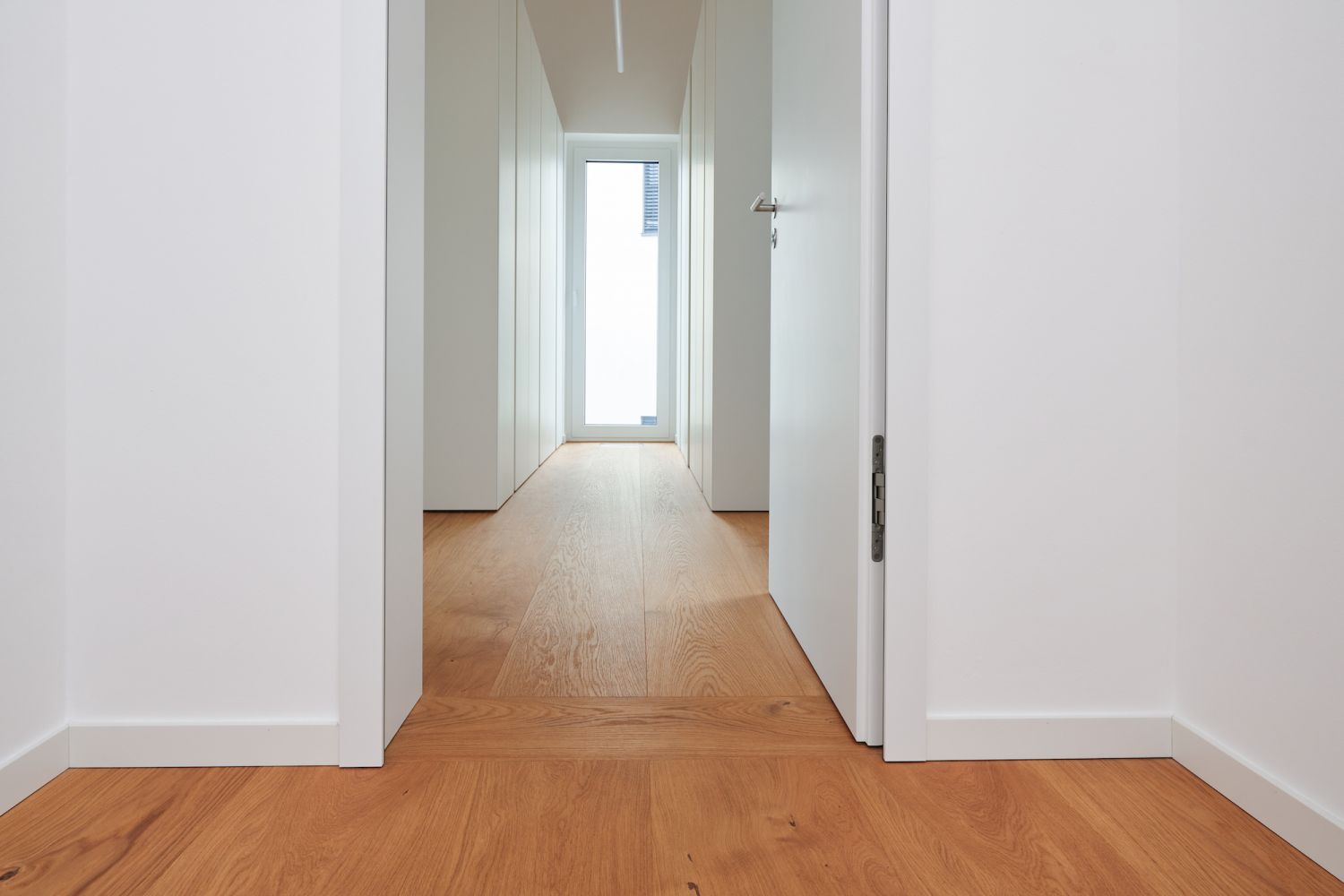 Blick durch einen weissen Flur mit offenem weissem Tuerblatt und Eiche Holzboden, am Ende ein Fenster mit Tageslicht.