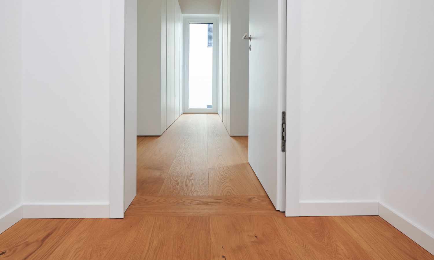 Blick durch einen weissen Flur mit offenem weissem Tuerblatt und Eiche Holzboden, am Ende ein Fenster mit Tageslicht.