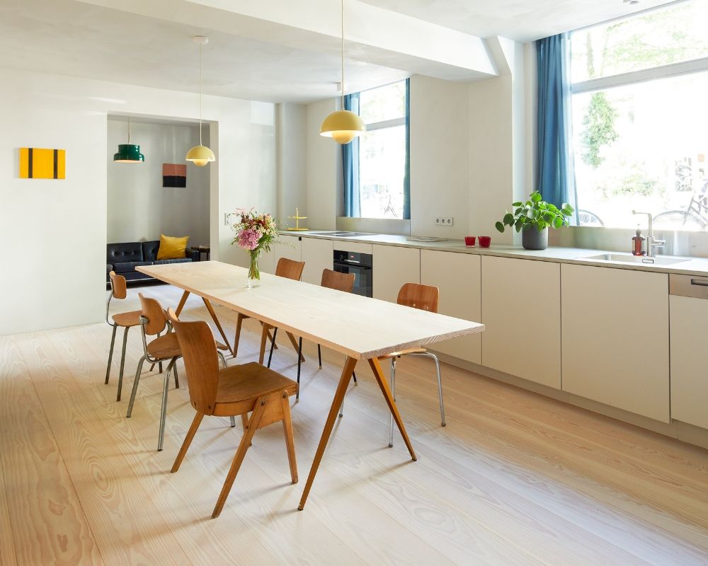 Coin repas moderne avec une longue table blanche; chaises en bois; cuisine avec évier; grandes fenêtres avec rideaux bleus
