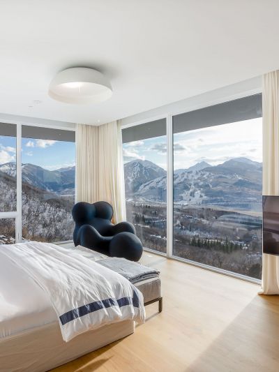 Schlafzimmer mit Eiche Massivholzdielen, grossem Bett, blau-weisser Bettwaesche, schwarzem Sessel und Panoramafenstern mit Blick auf schneebedeckte Berge.