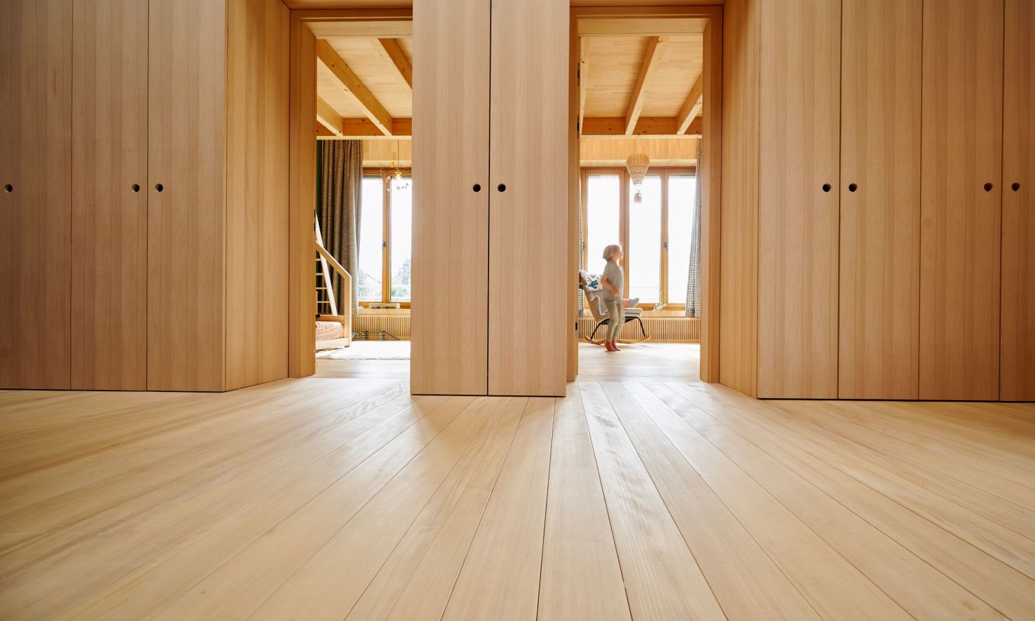 Vue a travers un couloir avec des sols et des murs en sapin blanc, avec une personne assise sur une chaise en arriere-plan.