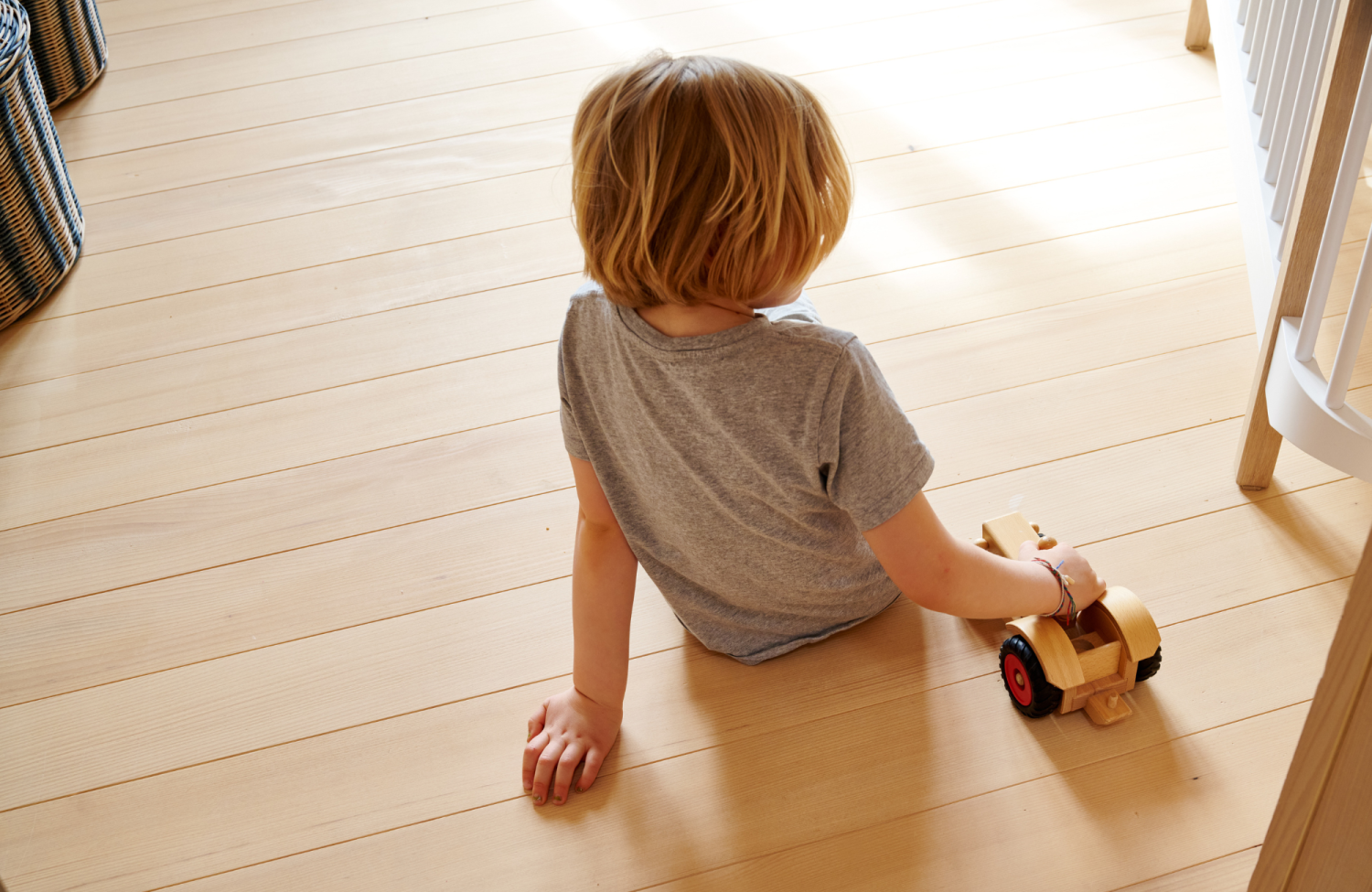 Enfant aux cheveux blonds et courts assis sur un plancher en sapin blanc tenant une petite voiture en bois aux roues rouges.