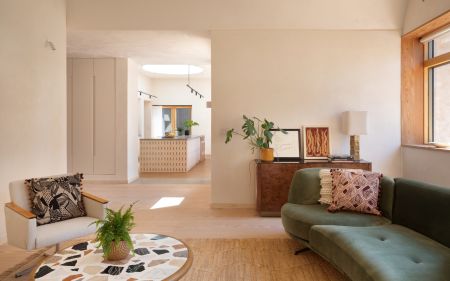 A living room with natural materials. Light brown fiber rug, table with a terrazzo top, dark green velvet sofa, and a light Douglas fir floor. Beyond, the view opens into the kitchen.