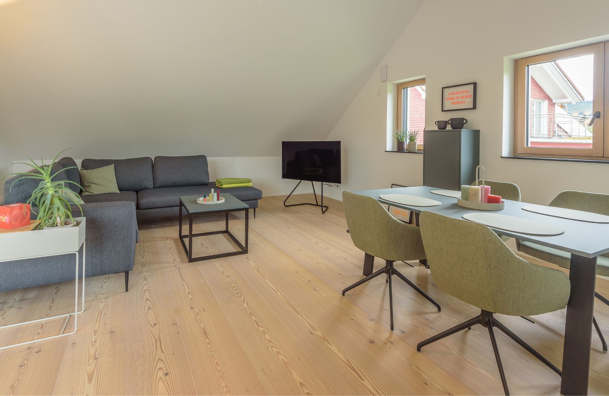 Salon avec canape d’angle gris, table basse noire, table a manger verte avec quatre chaises, televiseur sur meuble et deux fenetres.