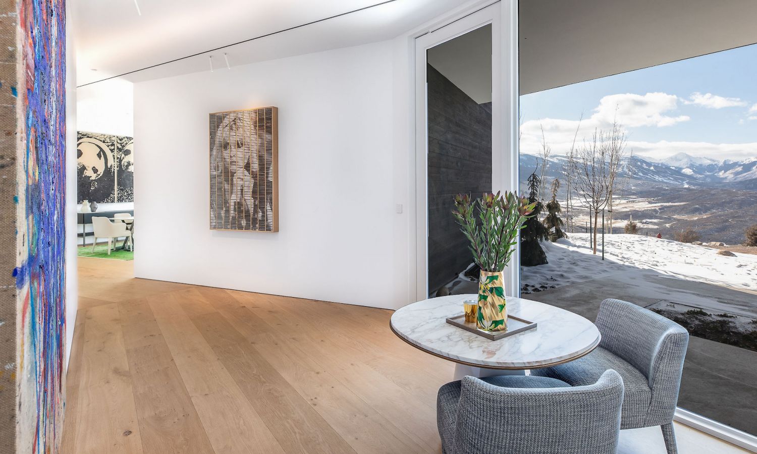 Modern living area with oak wood flooring, round marble coffee table, two grey armchairs and large window with mountain view.