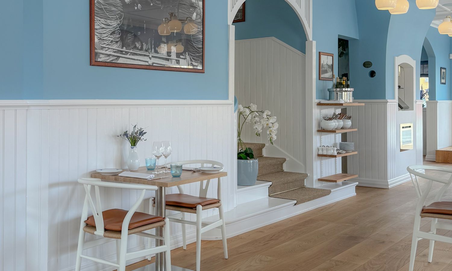 Light-blue dining area with white wood paneling, oak flooring, two tables with white chairs and brown seat cushions, a staircase, and a wall shelf with dishes.