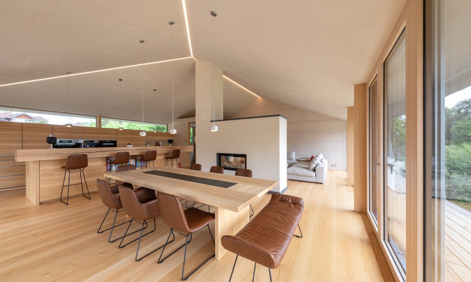 Salle à manger moderne avec une longue table en bois, des chaises en cuir marron, une cuisine ouverte et une grande fenêtre avec vue sur la verdure.