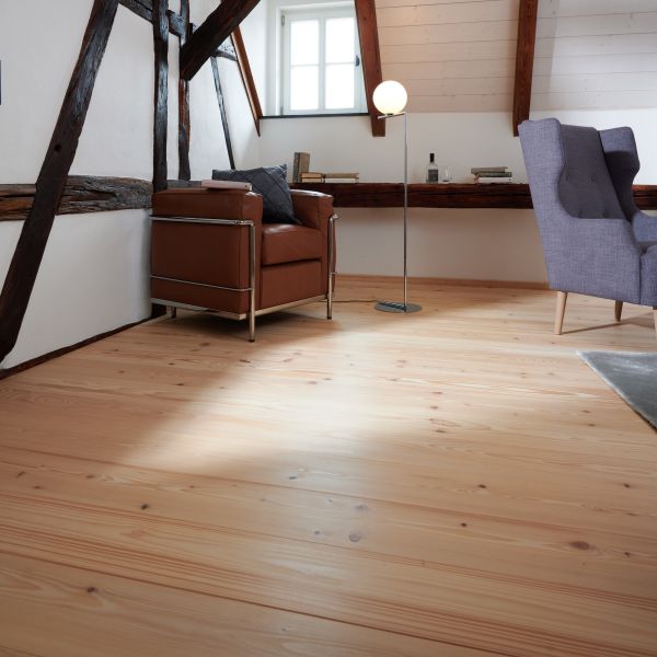 Helles Dachzimmer mit sichtbaren Holzbalken, braunem Ledersessel, Stehlampe mit runder Lampe und grauem Sessel.