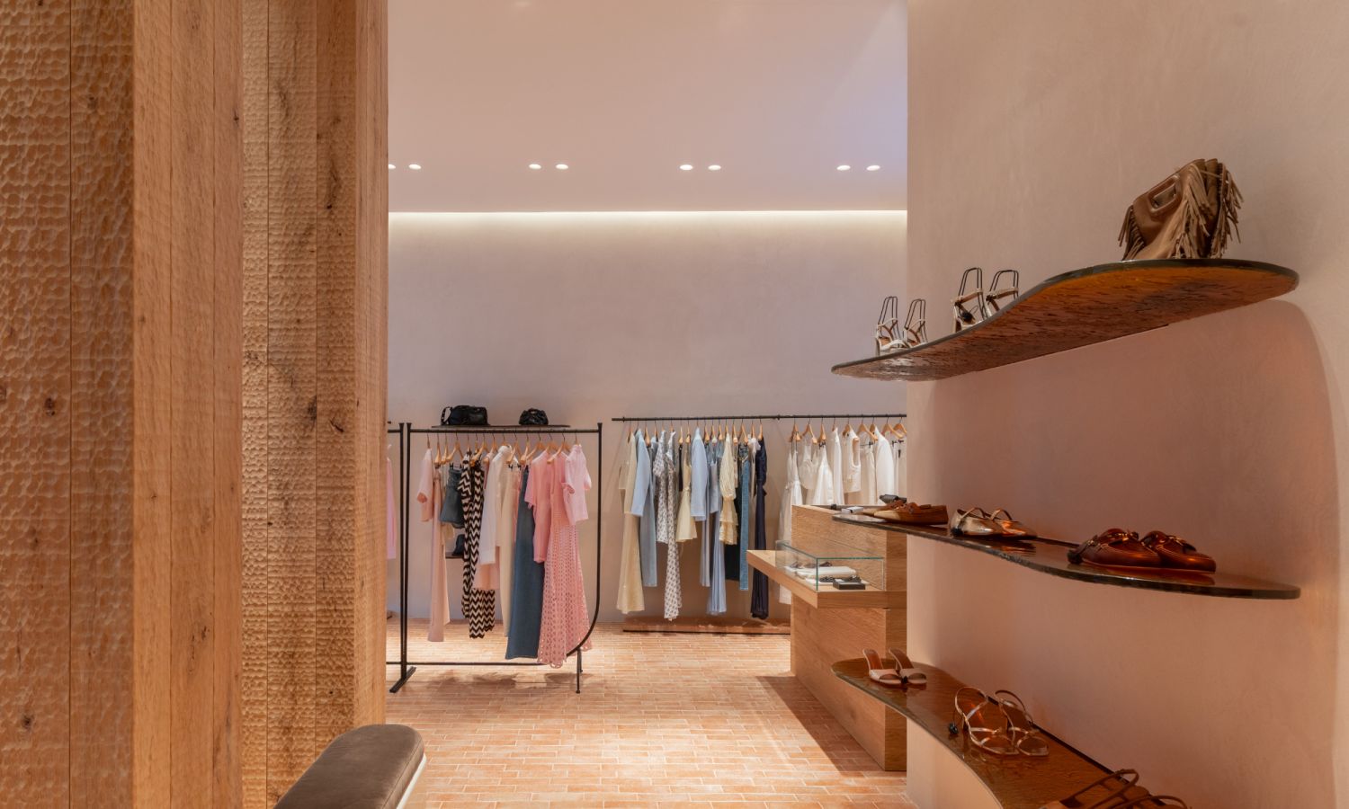 Vue intérieure de la boutique : à droite, une étagère aux formes arrondies présente des chaussures haut de gamme. À l’arrière-plan, des portants exposent des vêtements dans des tons clairs. À gauche, des panneaux muraux en bois texturés apportent chaleur