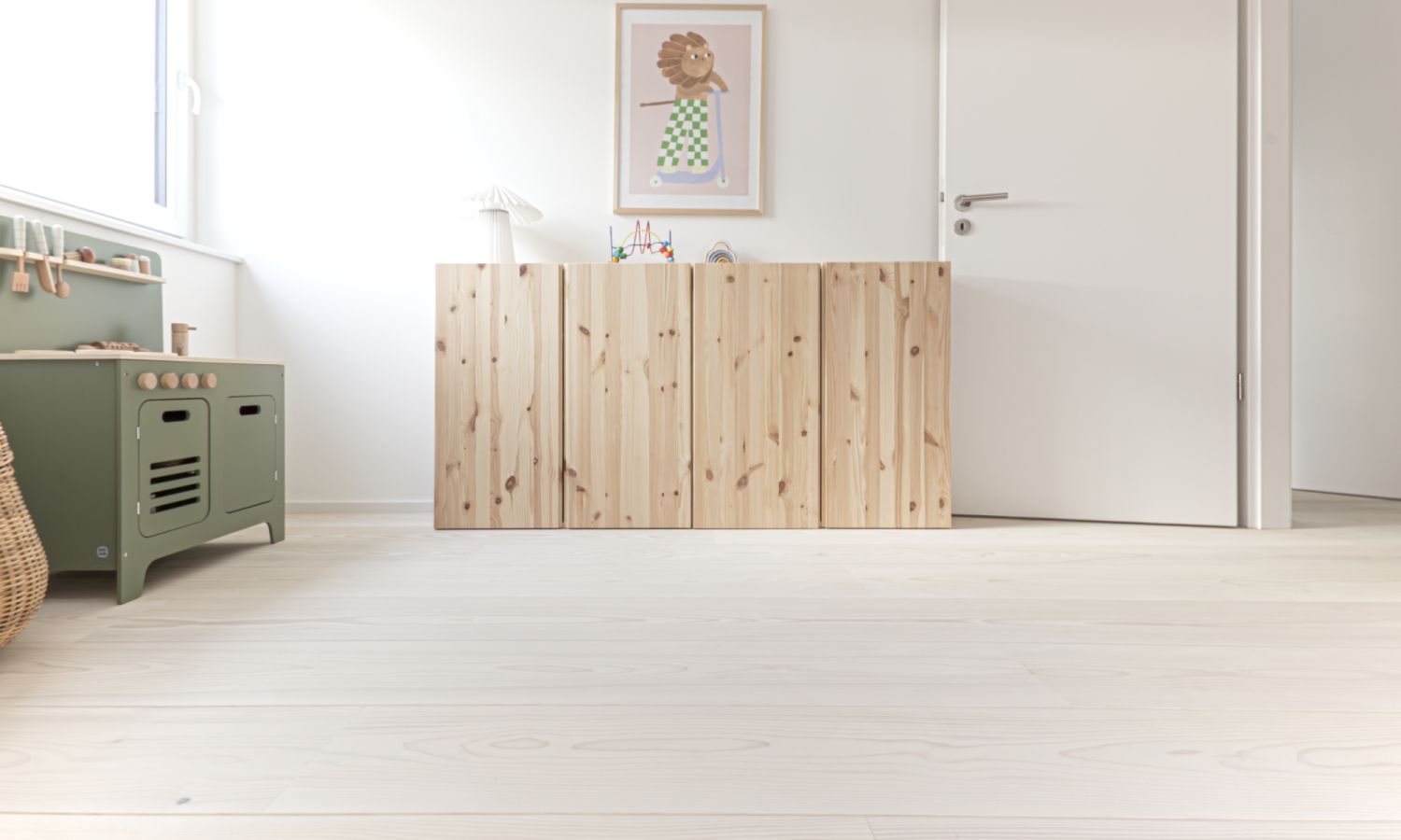 Heller Raum mit Weisstanne Holzboden, einem niedrigen Sideboard aus hellem Holz, einem Bild mit Blumenmotiv an der Wand und einer weissen Tuer.